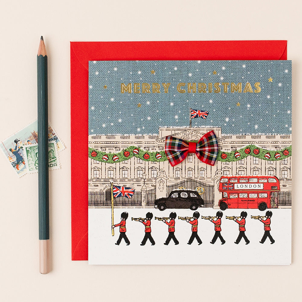 Christmas card with a festive design featuring a London scene, including a red double-decker bus and a marching band, with a red envelope.